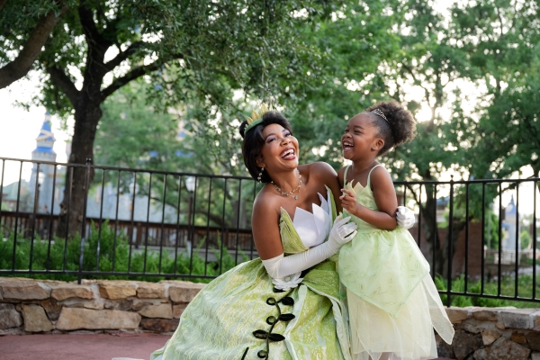 Princess Tiana in her green dress kneels beside a little girl also wearing a green dress.