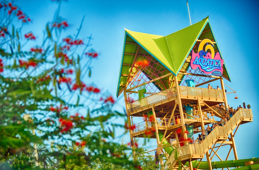 A wooden tower with a green roof and a large Aquatica sign with flowers and greenery in the foreground. 