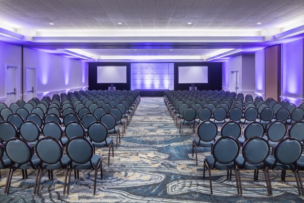 Rows of cushioned chairs on either side of an aisle facing the front of the room with projection screens and purple uplighting.