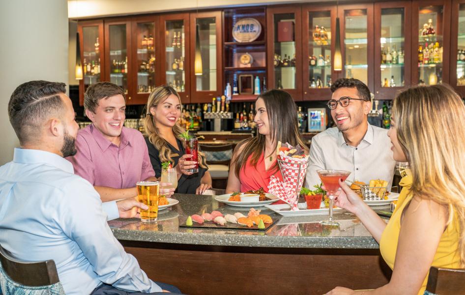 A group of adults sitting around a high top table sharing plates of food and toasting with cocktails. 