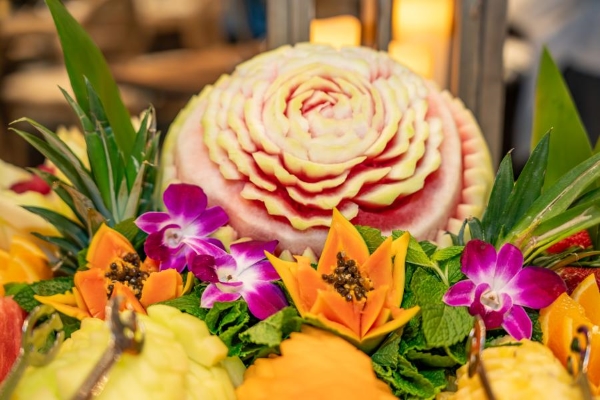 An intricately carved melon amidst a display of orchids, carved papayas, mint leaves, and pineapple fronds.