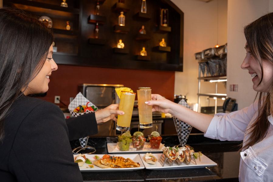 Two women toast with cocktails in highball glasses over plates of Mexican food.