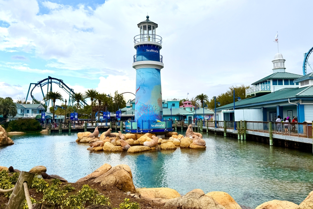 A lighthouse with the SeaWorld Orlando logo surrounded by statues of walruses and sea lions and a lagoon, with palm trees and a roller coaster in the background. 
