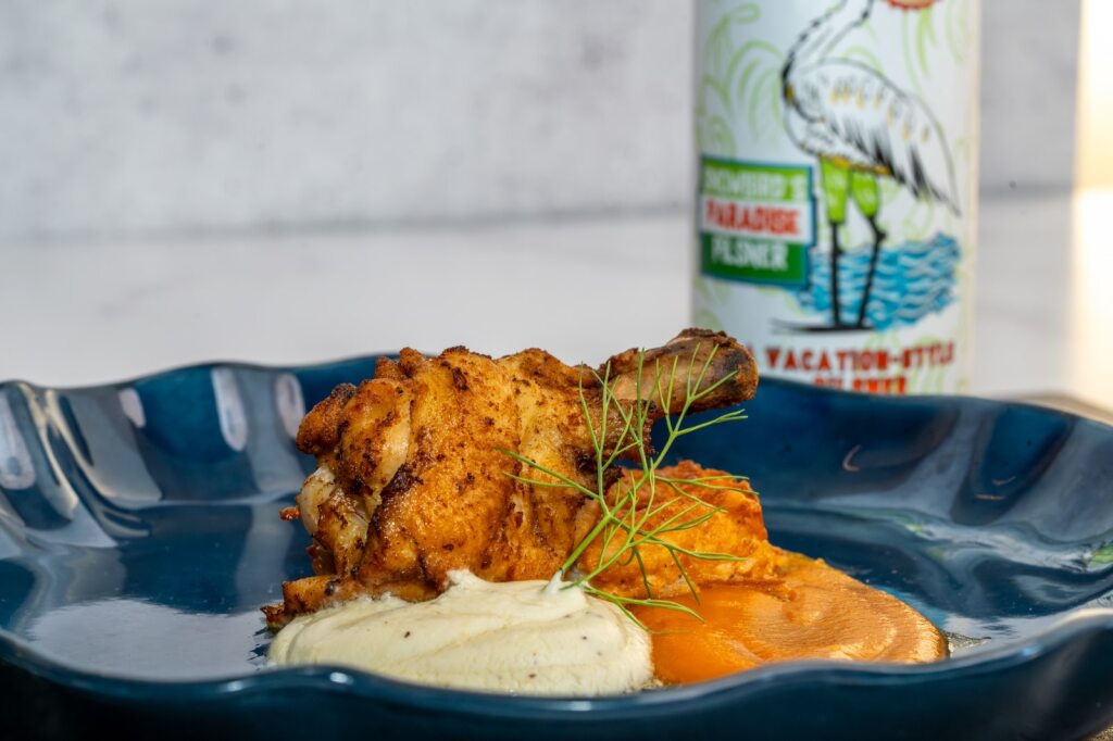 A close-up of a plate with a chicken leg, a light cream mousse, and an orange puree on a navy plate with a can of beer in the background.