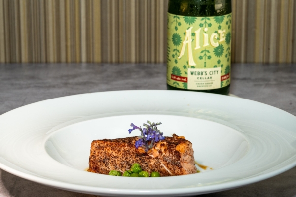 A piece of cooked salmon topped with edible flowers and served alongside green peas in a bowl beside a bottle with a green label that reads Alice Webb's City Cellar.