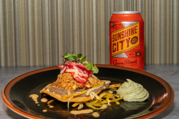A crispy tostada shell topped with shredded chicken, pink pickled onions, and microgreens with a dollop of pale green avocado crema, on a dark plate beside a bright orange can of Sunshine City beer.