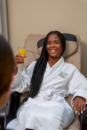 A woman in a white bathrobe sits in a pedicure chair with a mimosa in her hand. 