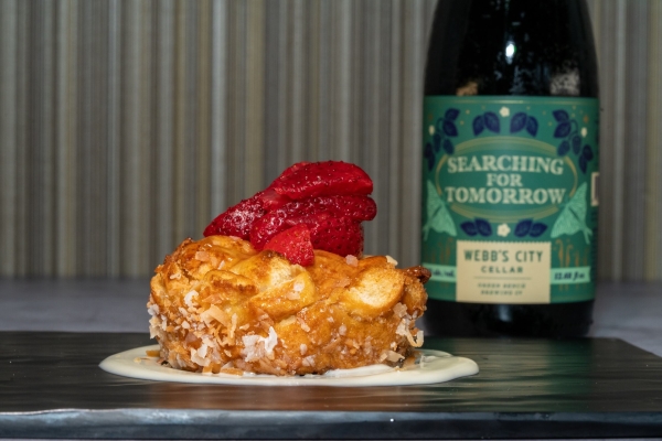 A beautiful round bread pudding dusted in coconut and topped with sliced strawberries beside a bottle with a green label that reads Searching for Tomorrow.