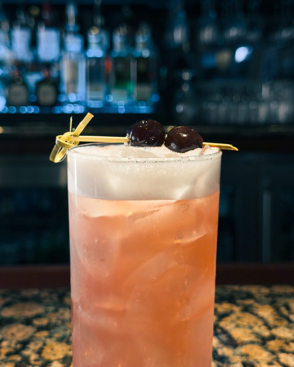 A close up of a pale pink cocktail with a black cherry garnish.