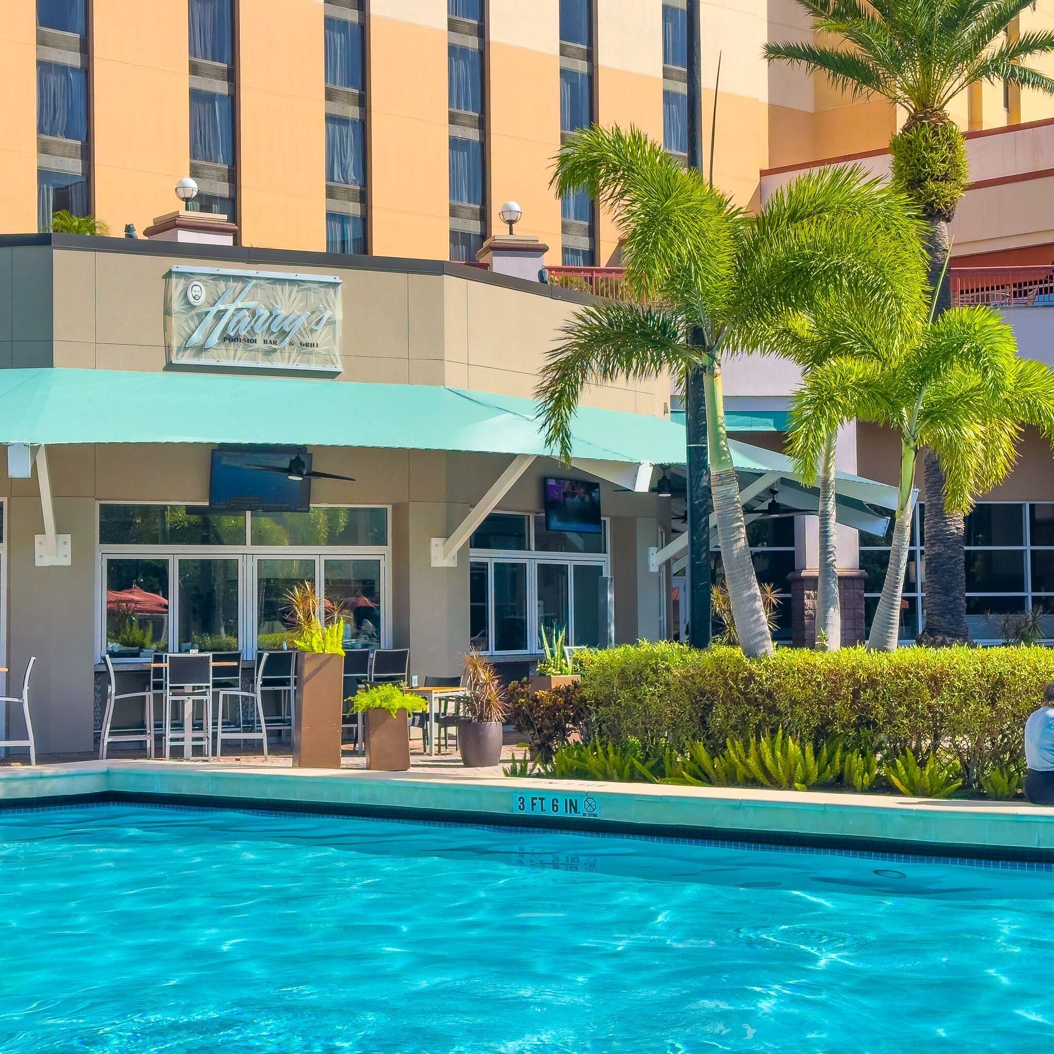 The front of a restaurant with a sign that reads Harry's Poolside Bar & Grill with outdoor seating beside a sparkling pool and palm trees.