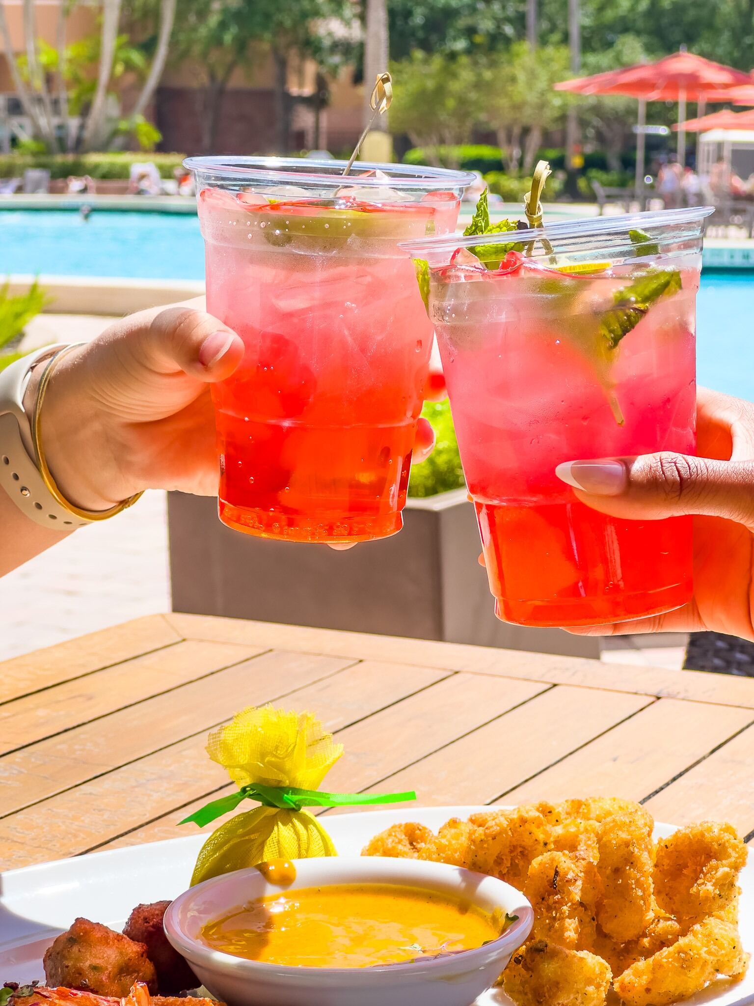 Two hands toast with bright pink cocktails over plates of food beside a pool. 