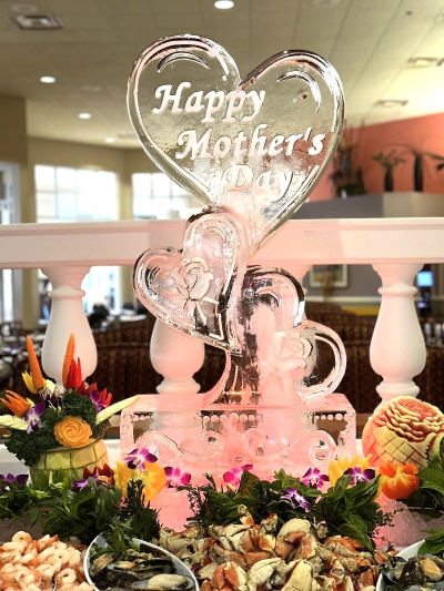 An ice sculpture centerpiece that says Happy Mother's Day with hearts surrounded by tropical fruit carvings and a seafood buffet.