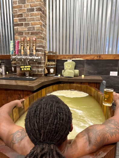 A man with dreadlocks sits in a wooden bath facing away from the camera looking towards a beer tap with a full pint glass in his hand. 