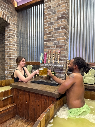 A man and woman in wooden beer tubs lean over a counter and cheers in front of a beer tap.