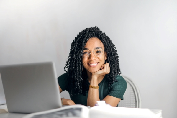 A young black woman with curly hair and glasses smiles at the camera sitting in front of an open laptop..
