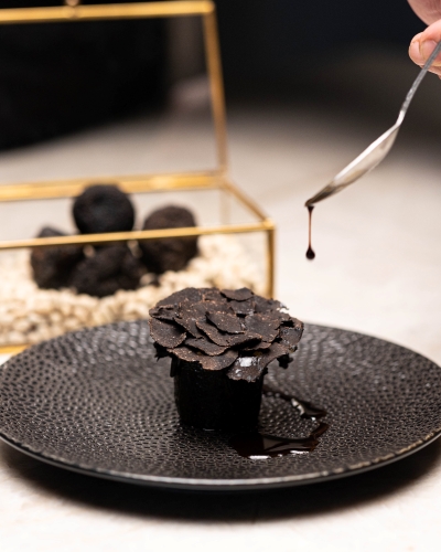 A spoon drips a finishing sauce onto a dark, flower-like food item on a black plate with a gold and glass box of black truffles in the background. Photo from Camille in Orlando, a Michelin Star restaurant.