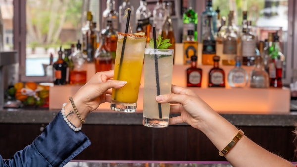 Two hands toasting cocktails in front of a glowing bar at Harry's Poolside Bar & Grill inside Rosen Centre.