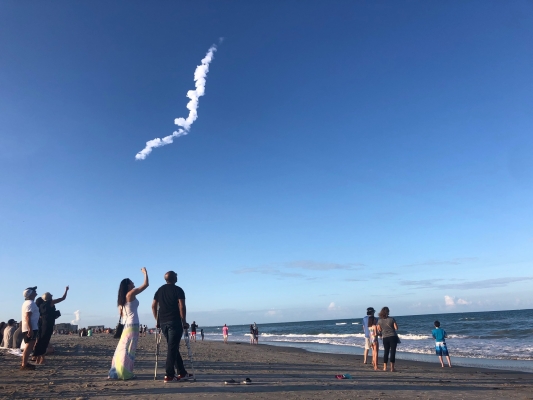 A beach with people standing and pointing at a clear blue sky with a trail of clouds from a rocket launch.