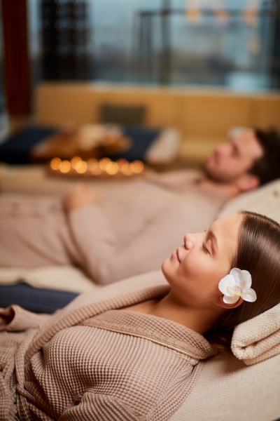 A man and a woman in robes lying on relaxing beds with candlelight with their eyes closed.