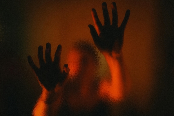 An out-of-focus figure in red light ominously presses their hands against a glass pane.
