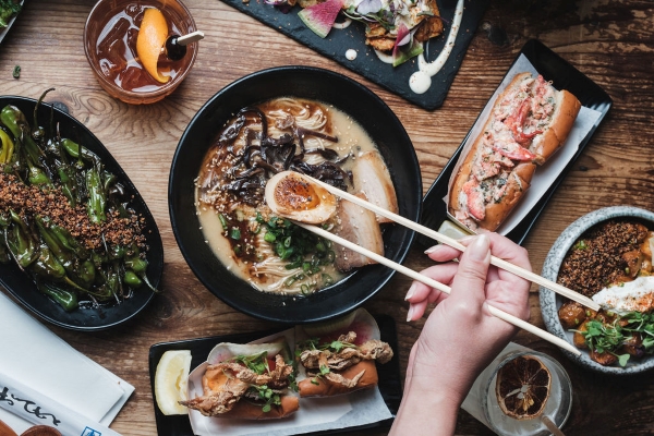 An overhead view of a hand holding chopsticks over a bowl of ramen and a table surrounded by delicious menu items.