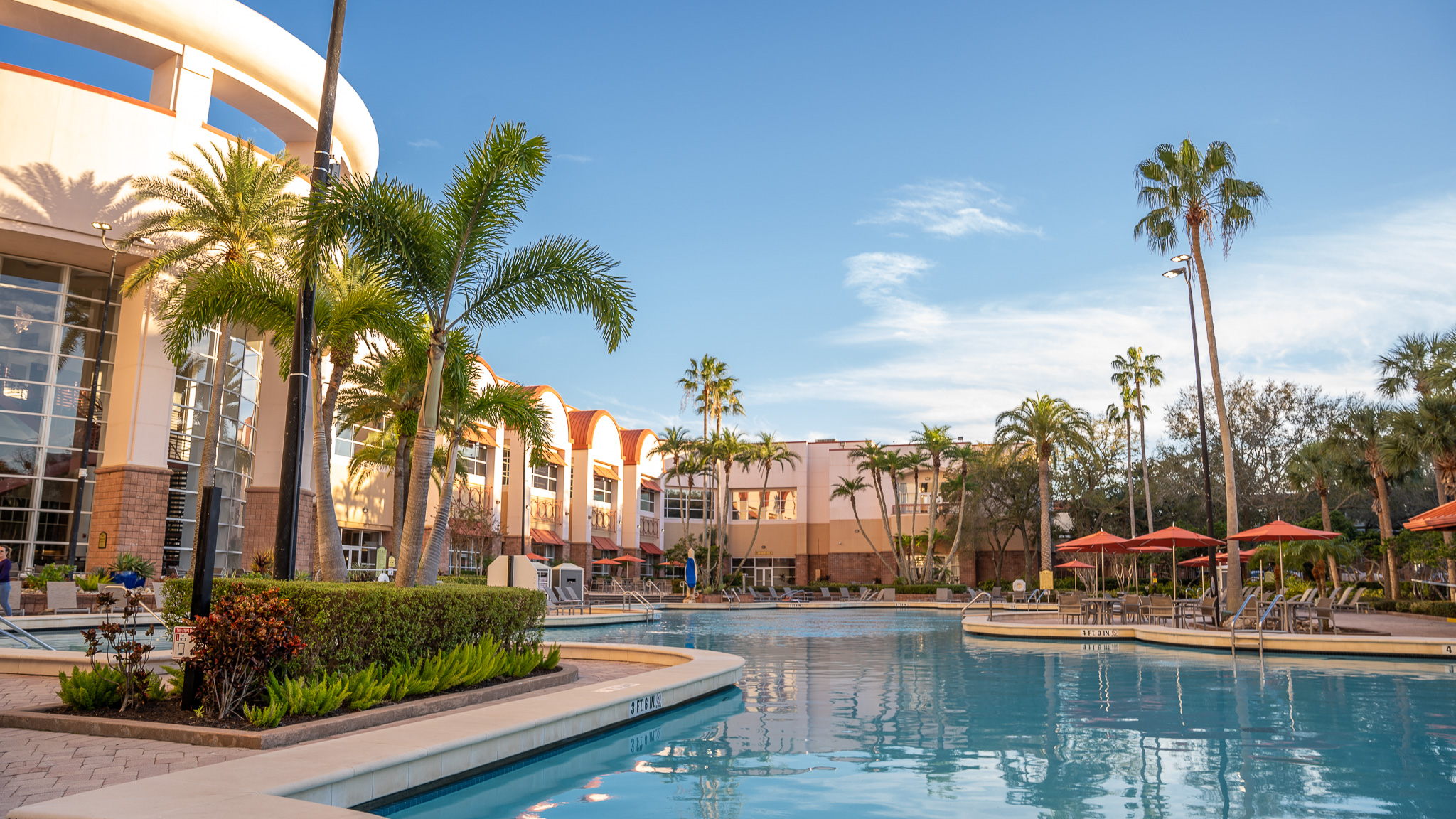 A multi-story beige and white Rosen Plaza Hotel building next to its bright blue swimming pool with palm trees around it on a sunny day. 