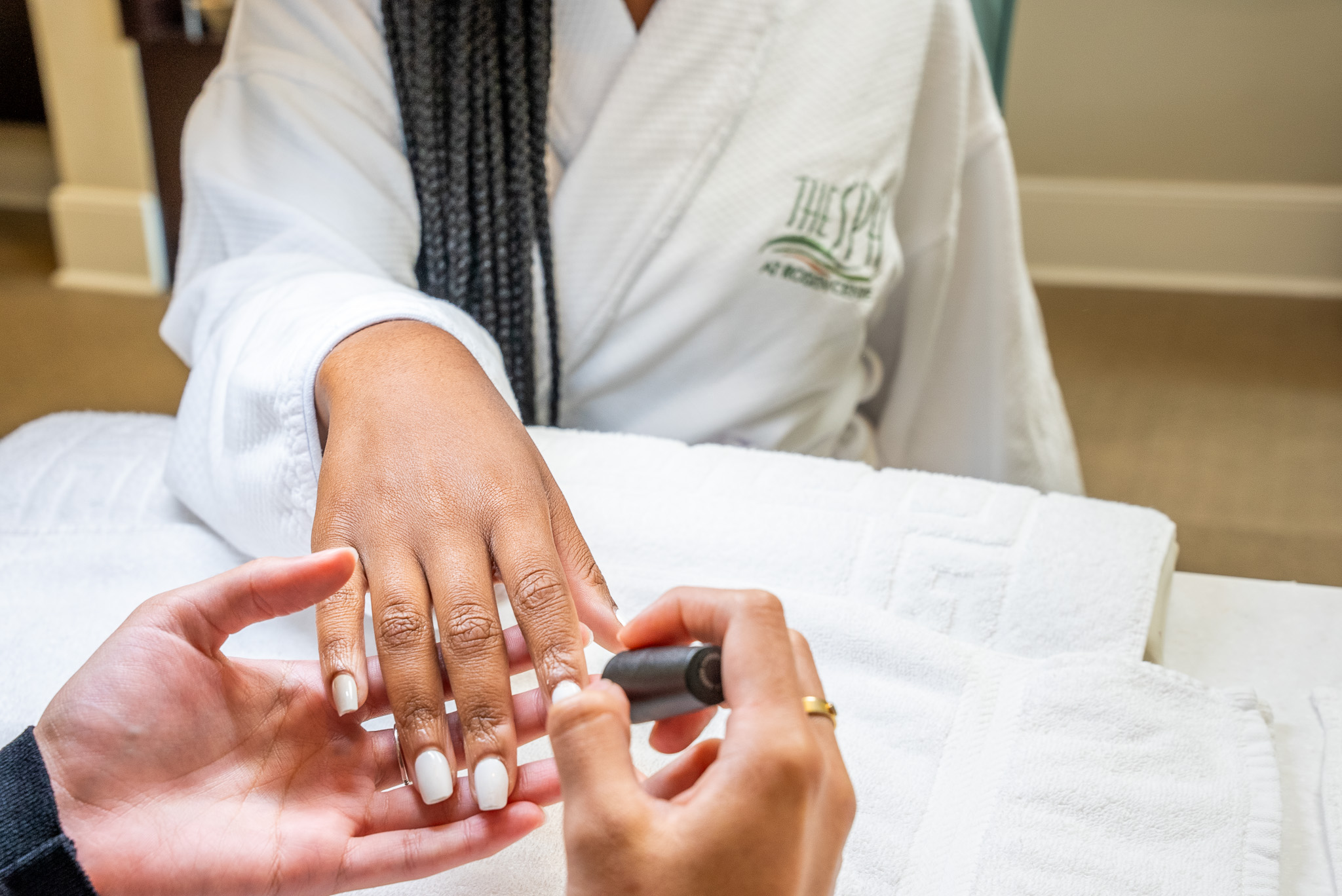 A woman’s hand is shown getting white nail polish applied by another pair of hands. The body of the woman getting her nails done is shown wearing a white robe with The Spa at Rosen Centre embroidery on it. 