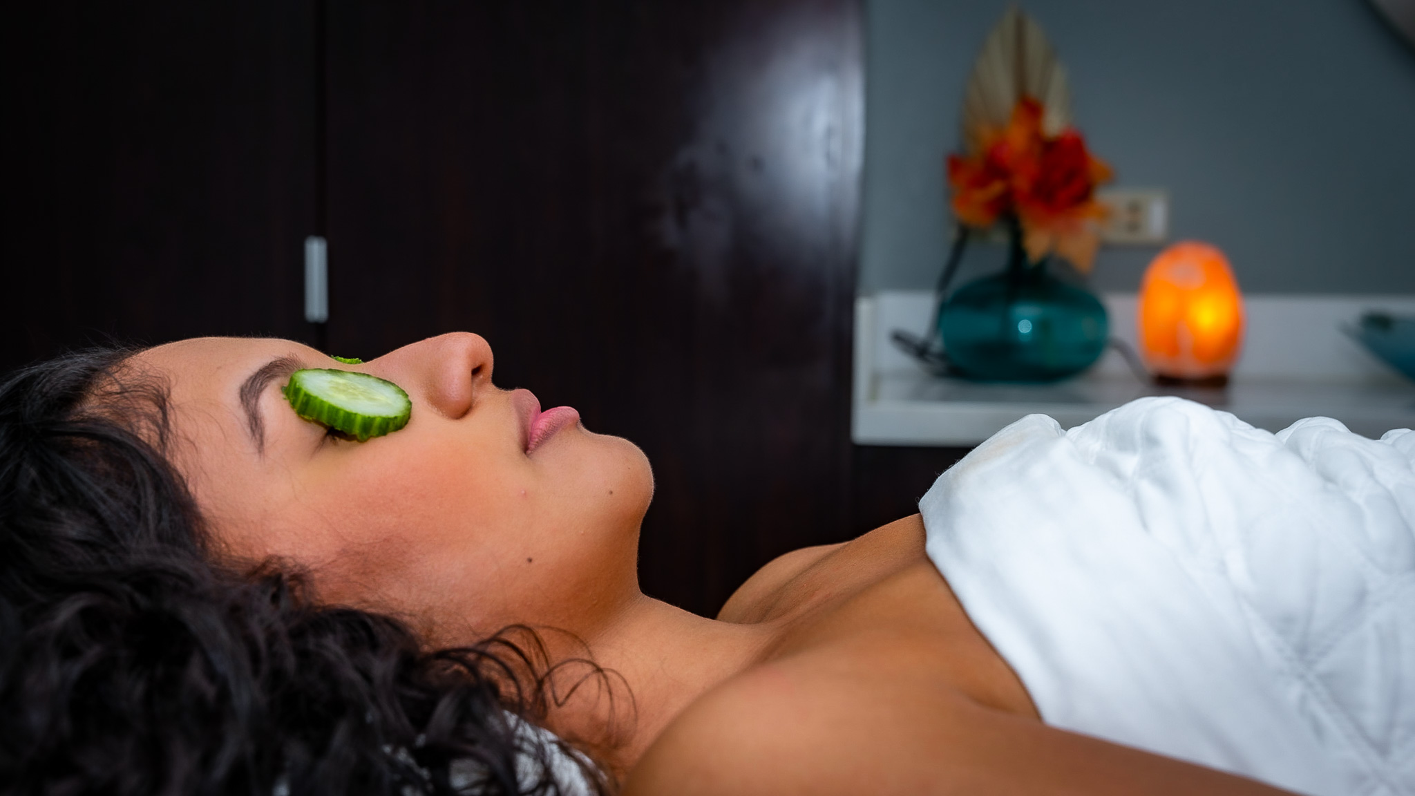 The profile of a woman with long dark hair is shown laying down with a cucumber on her eye. Her torso is seen wrapped in a white blanket. A red flower and lit up salt lamp is blurred in the background. 