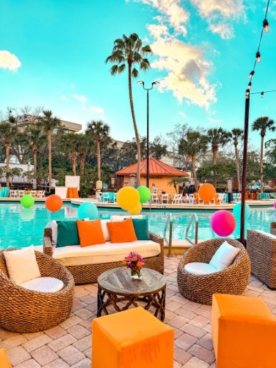 A poolside event with wicker lounge furniture, orange seating accents, and neon-colored balls floating in the pool.