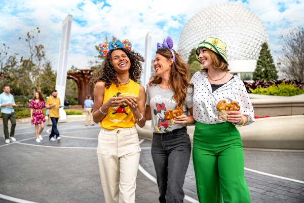 Three women in Mickey ears enjoy theme park food walking outside with Spaceship Earth behind them at EPCOT.