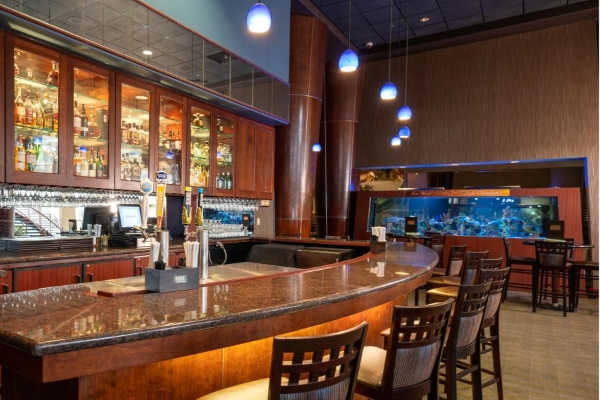 A bar space with a wall of glass-front cabinets showcasing bottles of liquor, high top chairs, and an aquarium in the background.