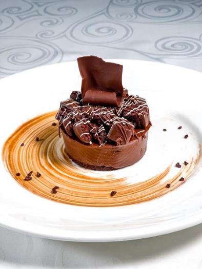 A small round chocolate cake garnished with drizzles of white chocolate and a chocolate ribbon on top.