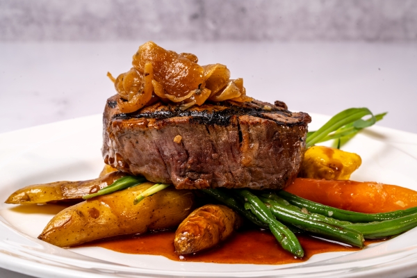 A tender steak topped with onions laying on a bed of roasted vegetables for Magical Dining 2026.