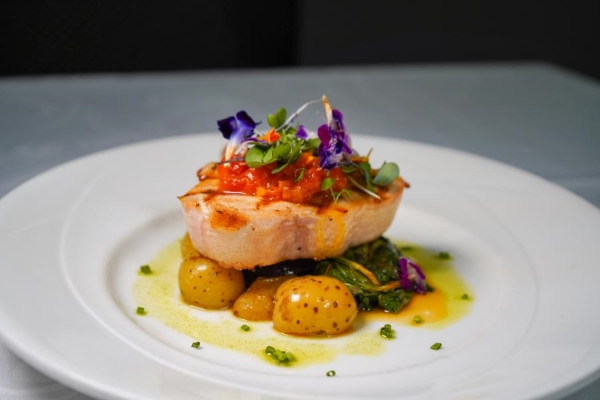 A beautiful piece of seared fish resting on potatoes and greens and topped with tomato jam, sprinkled greens, and edible flowers.
