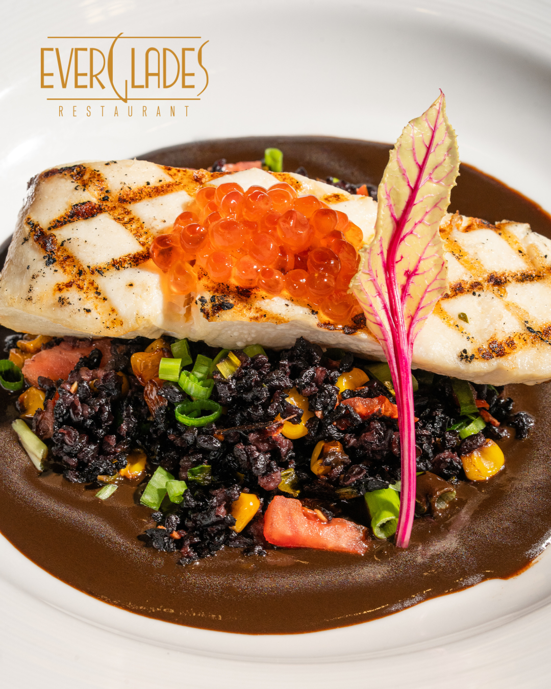 A seared filet of fish on a bed of black grains and colorful vegetables with the Everglades restaurant logo in the upper left corner.