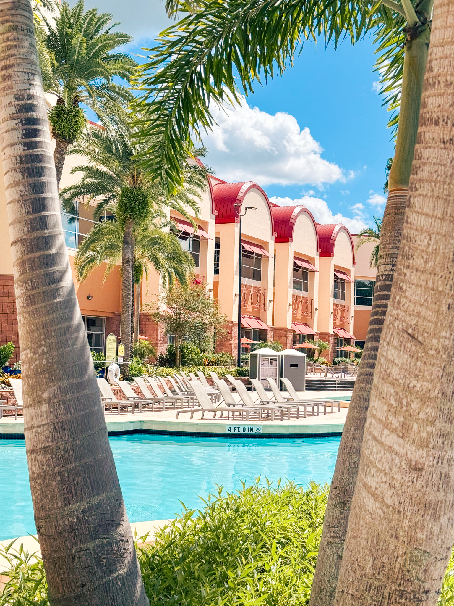 A two-story building with arched red roof peaks sits beside a pool surrounded by lounge chairs.