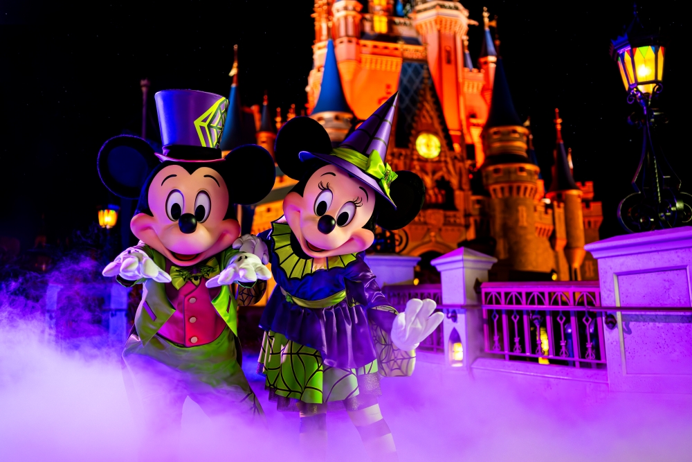 Mickey and Minnie in Halloween themed costumes standing in a purple haze in front of Cinderella's castle, glowing orange at night.