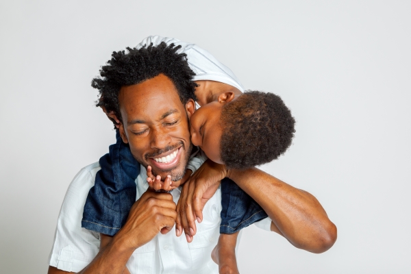 A boy in jeans and a white shirt sits on his dad’s shoulders, kissing him on the cheek as the dad smiles. Happy Father's Day.