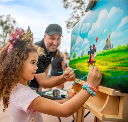 A girl in Minnie ears and her dad hold paintbrushes to a canvas painting of Mickey and Minnie sitting on a bench in a field of flowers. 