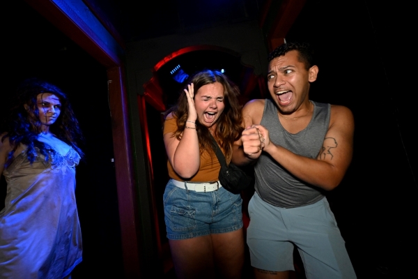 A man and woman screaming cling to each other in a dark room with a monster crawling out of the shadows beside them.