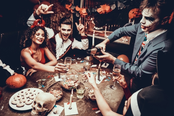 A group of adults in Halloween costumes sit around a table as a man pours Champagne. 