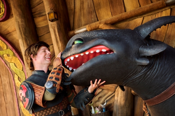 A young man in Viking uniform pets the nose and chin of a black smiling dragon.