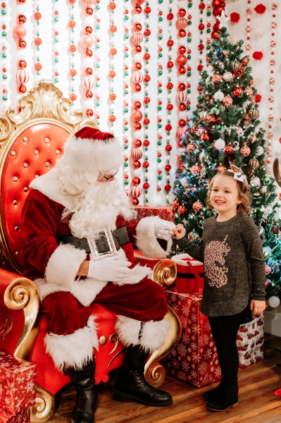 Santa in a big red chair greets a little girl in front of a wall draped in ornaments and a Christmas tree at the Winter Wonderland at Wekiva Island. 