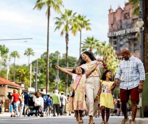 A family with two daughters smiles and walks along a street with Hollywood Tower of Terror and palm trees behind them.
