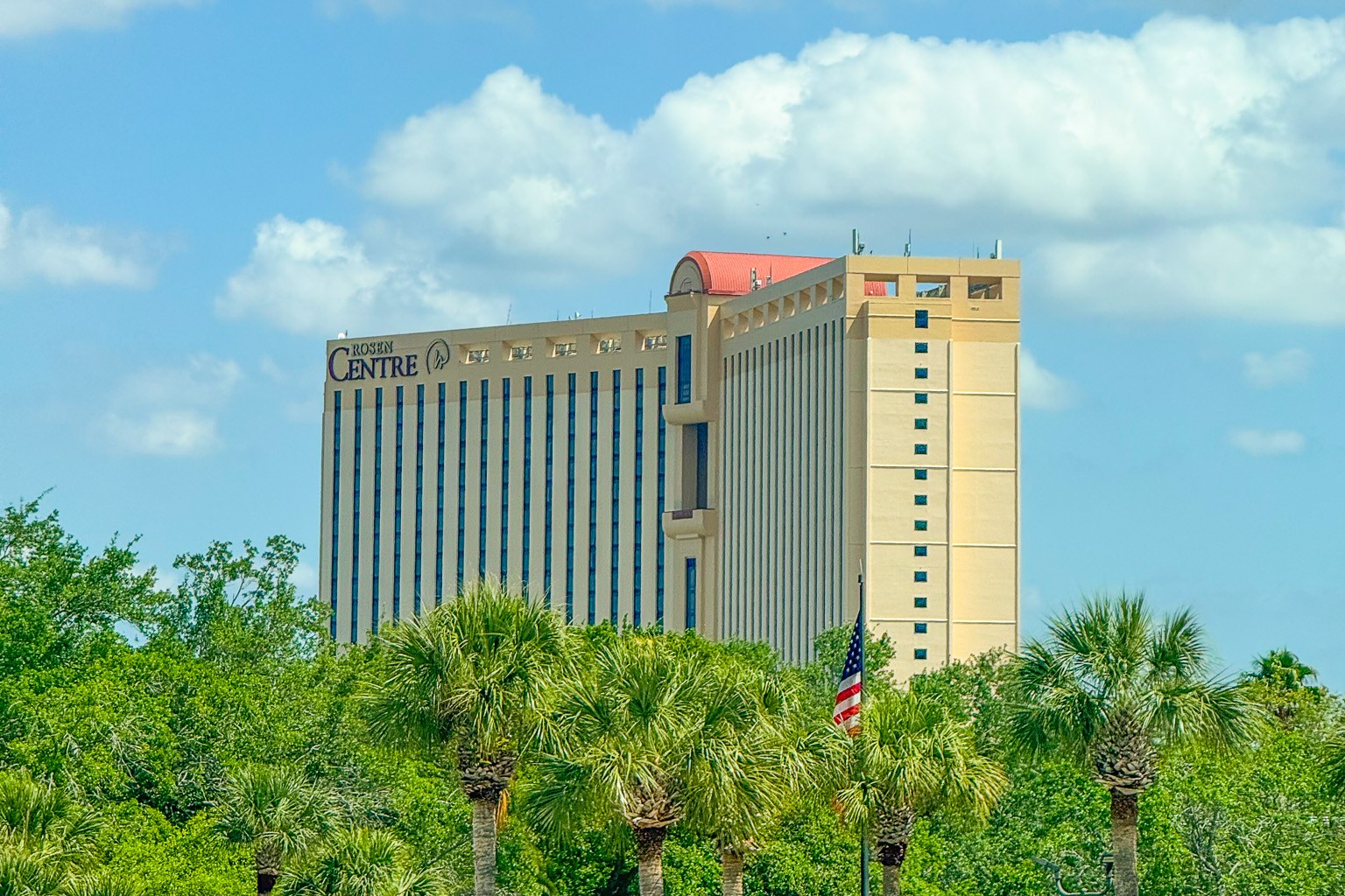 A tall hotel building, Rosen Centre, rises above the treetops against a blue and cloudy sky. 