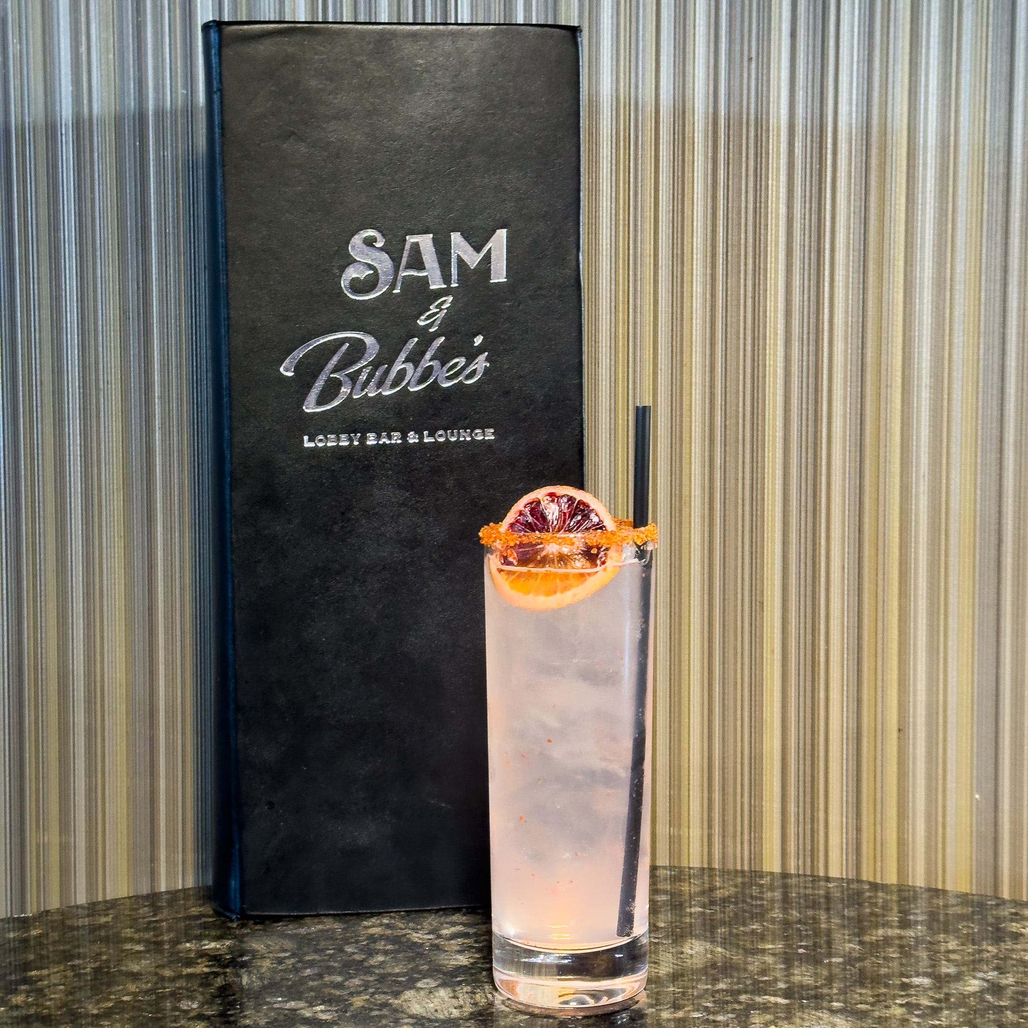 A pale pink cocktail with a dried citrus garnish in front of a Sam and Bubbes Menu