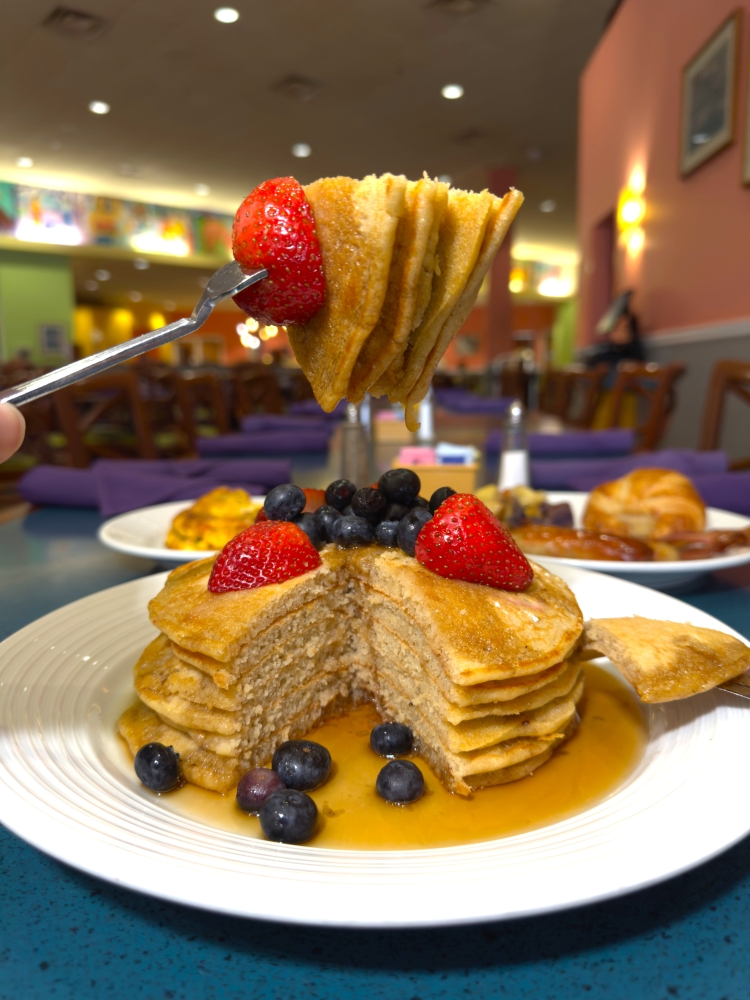 A fork holds a bite of stacked pancakes in front of a plate of pancakes topped in syrup and berries. 
