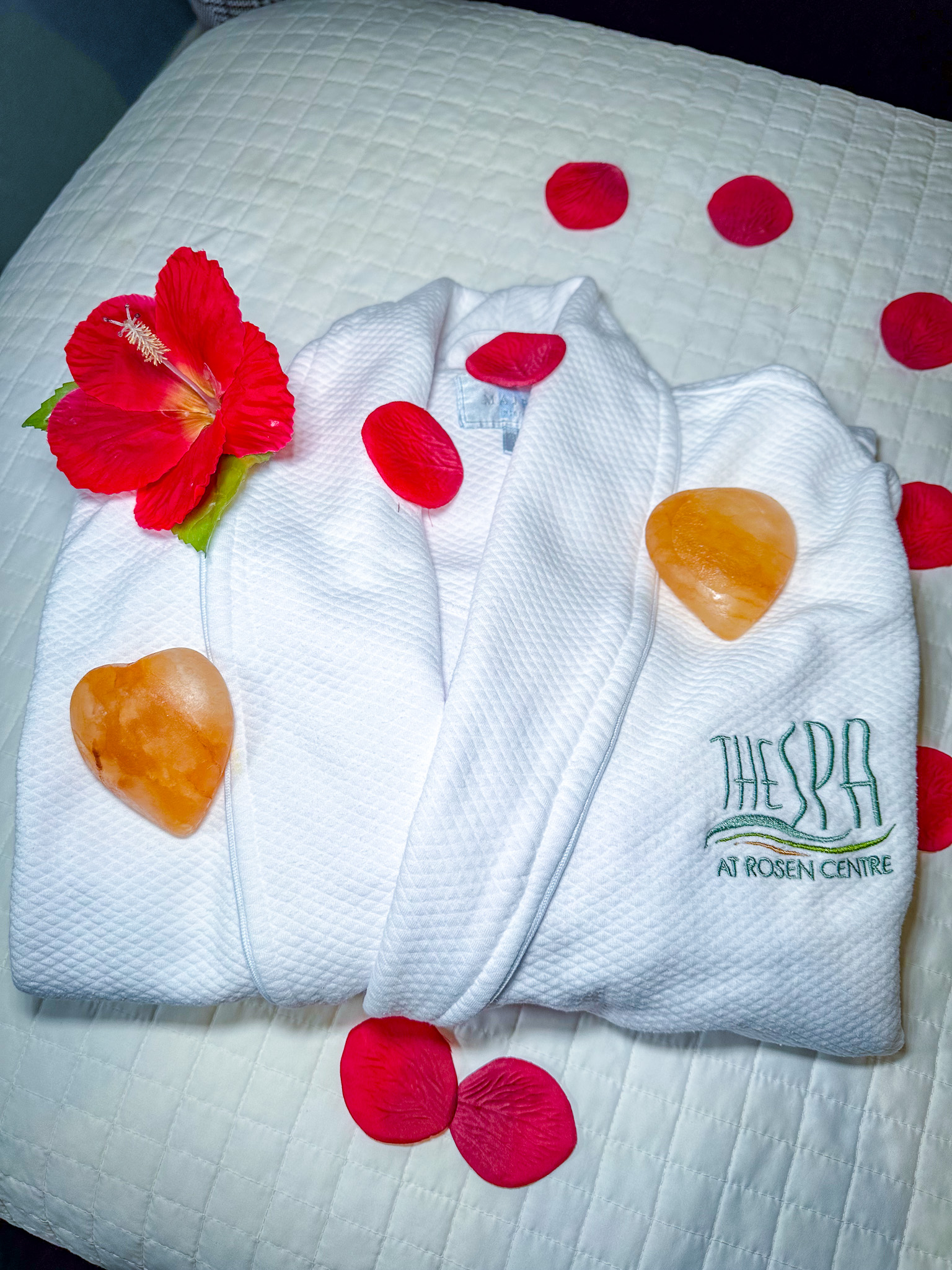 A white spa robe with the words "THE SPA AT ROSEN CENTRE" embroidered on it in seafoam green and gold thread with a red flower and two amber-colored stones rests on a white textured blanket. Red flower petals are scattered around it.