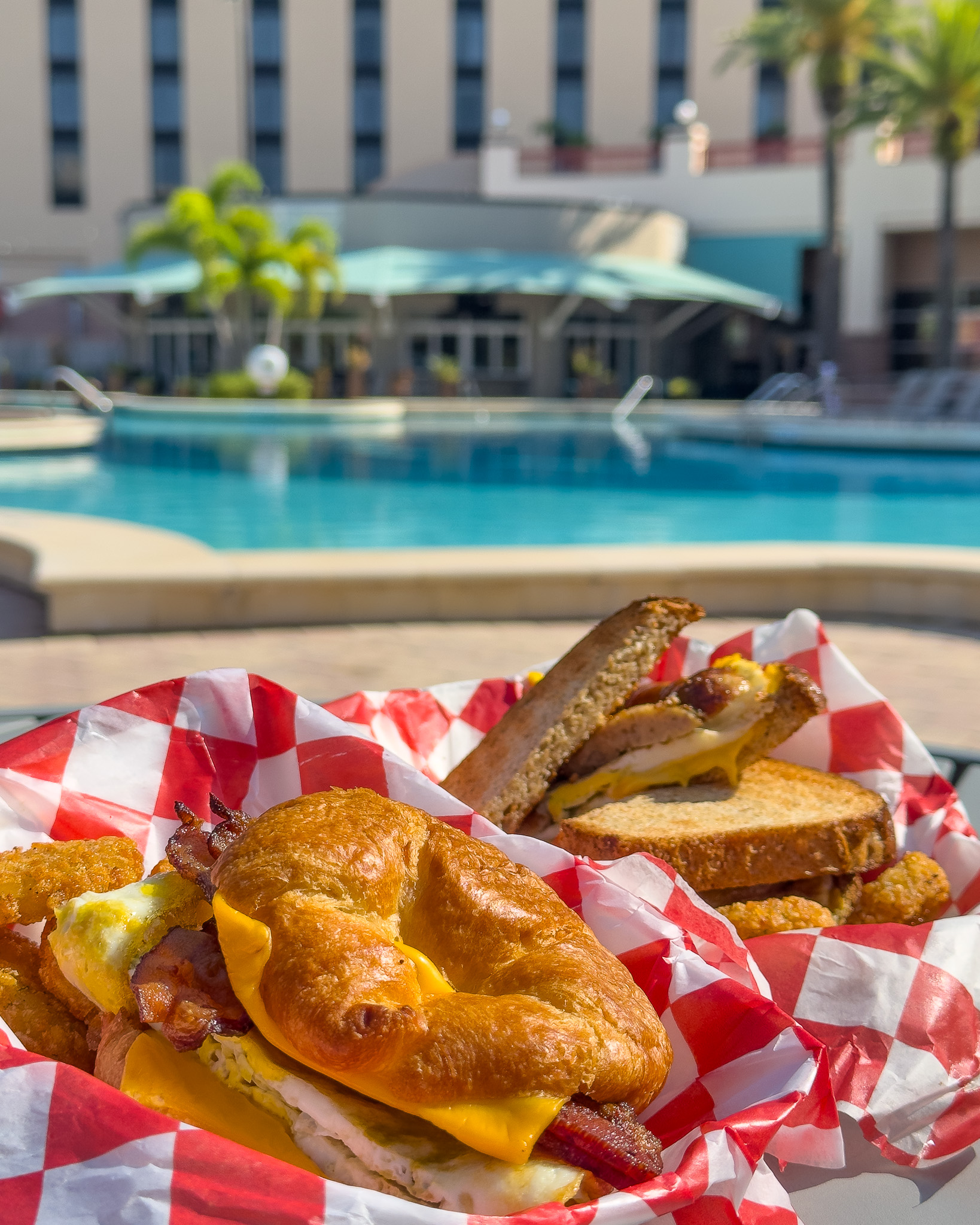 Two breakfast sandwiches in baskets with red and white checked paper on a table in front of a beautiful pool. 
