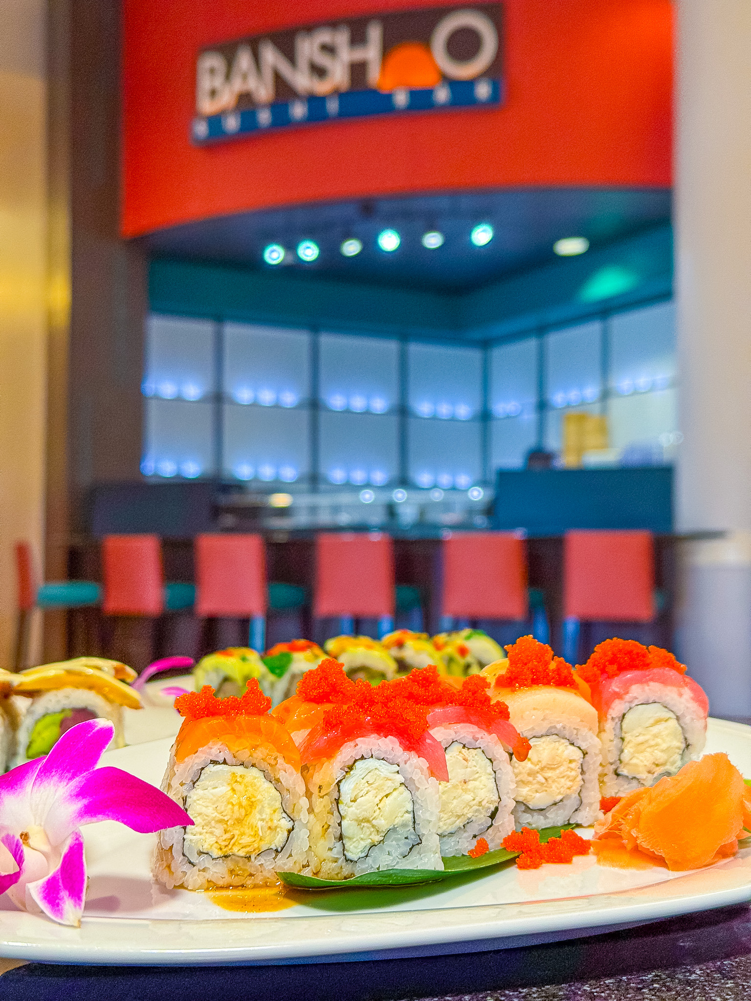 Colorful plates of sushi in the foreground with red leather high top stools and the sign for Banshoo Sushi in the background. 